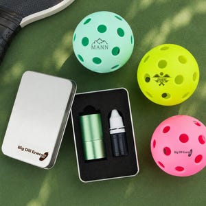Op de afbeelding: Drie geperforeerde pickleballen in mintgroen, felgeel en neonroze, elk met aangepaste tekst en afbeeldingen. Een rechthoekige zilveren metalen behuizing met de tekst Big Dill Energy bevat een groene cilindrische buis en een klein zwart druppelflesje.