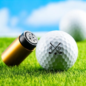Könnte beinhalten: Ein weißer Golfball mit einem schwarzen Logo, das "RG" mit zwei gekreuzten Golfschlägern zeigt. Das Logo ist mittig auf dem Ball platziert. Der Ball liegt auf grünem Gras.