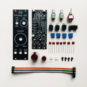 May include: An assortment of electronic components, including circuit boards, potentiometers, and capacitors. The components are arranged on a white surface. The text "WILD SYNC FATMOOG UCF" is visible on a black panel. Other components include a multi-colored ribbon cable.