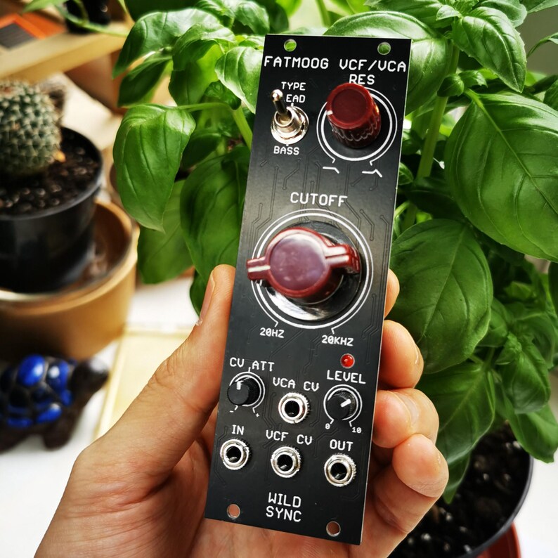Puede incluir: Un m&oacute;dulo electr&oacute;nico negro con perillas rojas y texto blanco, incluyendo "FATMOOG UCF/UCA RES" y "CUTOFF". El m&oacute;dulo tiene varias entradas y salidas, y el texto "WILD SYNC" est&aacute; en la parte inferior. El m&oacute;dulo se sostiene frente a follaje verde.