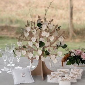Peut inclure: Un arbre en bois en forme d'arbre de mariage avec des étiquettes en forme de cœur blanches suspendues aux branches. Les étiquettes sont décorées de noms et de messages écrits à la main. L'arbre est entouré d'enveloppes blanches avec des nœuds en ficelle et un oreiller blanc avec un motif en forme de cœur.