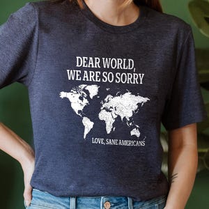 May include: Navy blue t-shirt with the text "DEAR WORLD, WE ARE SO SORRY" and a white world map graphic. Below the graphic, the text reads "LOVE, SANE AMERICANS". The shirt is part of the Double Mood Collection.