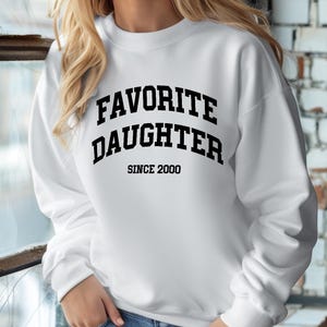 May include: White sweatshirt with the text "FAVORITE DAUGHTER SINCE 2000" in black block letters. The sweatshirt is long-sleeved and has a crew neck. The model is wearing blue jeans.