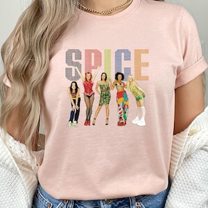 May include: A light pink t-shirt featuring the word "SPICE" in a colorful, pixelated font. Below the text are illustrations of five women in various outfits. The shirt has short sleeves and a crew neck.