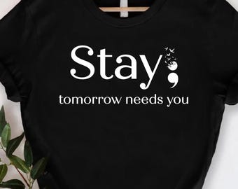 May include: Black t-shirt with the words "Stay; tomorrow needs you" in white. The semicolon is decorated with small bird silhouettes. The shirt is hanging on a wooden hanger against a white background.