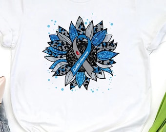 Puede incluir: Camiseta blanca con un diseño de girasol. El girasol tiene pétalos azules y grises, una cinta azul con "Diabetes Awareness" y un estampado de leopardo. La camiseta tiene mangas cortas y cuello redondo.