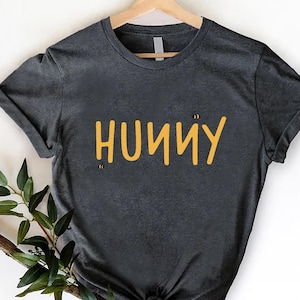 以下が含まれることがあります： 黄色で「HUNNY」の文字と2匹の小さな蜂のイラストが描かれた濃い灰色のTシャツ。木製のハンガーに掛けられ、裾に結び目があります。緑の小枝が横に置かれています。