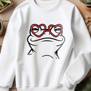 May include: White sweatshirt featuring a cartoon frog face wearing red heart-shaped glasses. The frog is drawn in black, with a smiling expression. The sweatshirt is a casual style, suitable for everyday wear.