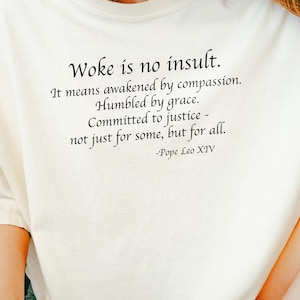 以下が含まれることがあります： 「Woke is no insult.」のテキストと教皇レオ14世の引用句が黒でプリントされたクリーム色のTシャツ。柔らかい素材で作られています。