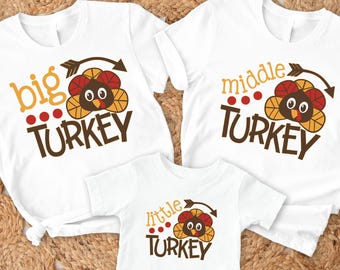 May include: Three white t-shirts with turkey-themed graphics. The shirts read "big turkey", "middle turkey", and "little turkey" in brown and orange lettering. Each shirt features a cartoon turkey design with an arrow.