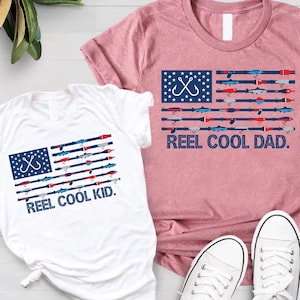 May include: Two t-shirts with a fishing theme. One white t-shirt has a graphic of a fishing hook and fishing rods arranged like the American flag, with the text "REEL COOL KID." The other is a pink t-shirt with the same design and the text "REEL COOL DAD."