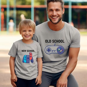 Op de afbeelding: Twee grijze t-shirts met bijpassende ontwerpen. E&eacute;n shirt zegt "OLD SCHOOL" met een controller-afbeelding. De andere zegt "NEW SCHOOL" met een gameconsole-afbeelding. De shirts worden gedragen door een man en een kind.
