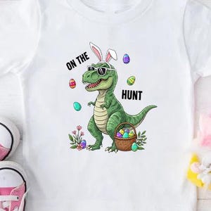 May include: White t-shirt with a graphic of a green dinosaur wearing sunglasses and bunny ears, holding a basket of Easter eggs. The text "ON THE HUNT" is above and below the dinosaur. Pink sneakers and Easter eggs are also in the image.