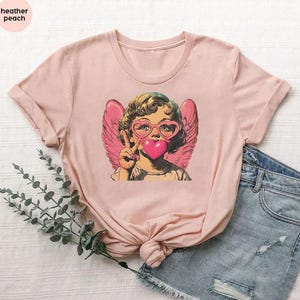 May include: A pink t-shirt with a graphic of a cherub wearing heart-shaped glasses and blowing a pink heart bubble. The cherub has blonde hair and is wearing a white shirt. The t-shirt has a heather peach color.