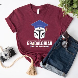 以下が含まれることがあります： マルーンのTシャツに、マンダロリアンのヘルメットの上に卒業帽をかぶったグラフィックがプリントされています。グラフィックの下には「The Gradalorian This is the Way」というテキストがプリントされています。