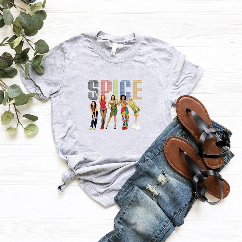 K&ouml;nnte beinhalten: Ein hellgraues T-Shirt mit dem Wort "SPICE" in einer bunten, pixeligen Schriftart. Darunter befinden sich Illustrationen von f&uuml;nf Frauen. Das Shirt wird mit einer blauen Jeans und braunen Sandalen kombiniert.