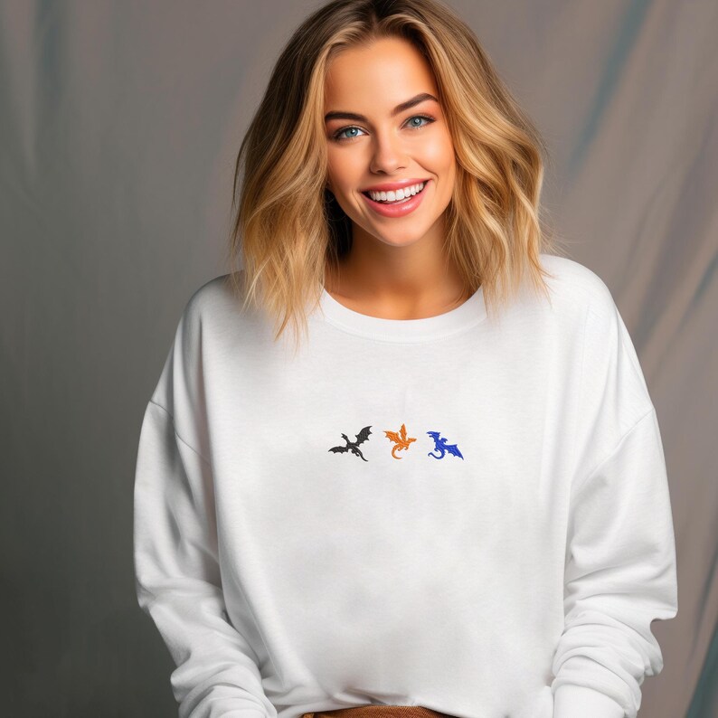 May include: White sweatshirt with three embroidered dragon silhouettes in black, orange, and blue. The crew neck sweatshirt is made of a soft material and is suitable for casual wear.