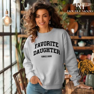 May include: A light gray sweatshirt with the words "FAVORITE DAUGHTER" in bold black letters, with "SINCE 2000" below. The sweatshirt is part of the Double Mood Collection.
