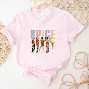 K&ouml;nnte beinhalten: Ein hellrosa T-Shirt mit dem Wort "SPICE" in einer bunten, pixeligen Schrift. Darunter befinden sich Illustrationen von f&uuml;nf Frauen in verschiedenen Outfits. Das T-Shirt ist unten geknotet.