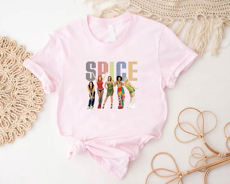 K&ouml;nnte beinhalten: Ein hellrosa T-Shirt mit dem Wort "SPICE" in einer bunten, pixeligen Schrift. Darunter befinden sich Illustrationen von f&uuml;nf Frauen in verschiedenen Outfits. Das T-Shirt ist unten geknotet.