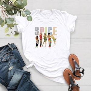 K&ouml;nnte beinhalten: Wei&szlig;es T-Shirt mit einem farbenfrohen Grafikdesign der Spice Girls und dem Wort "SPICE" dar&uuml;ber. Das Design zeigt die f&uuml;nf Mitglieder in verschiedenen Outfits. Das Shirt wird mit blauen Jeans und braunen Sandalen pr&auml;sentiert.