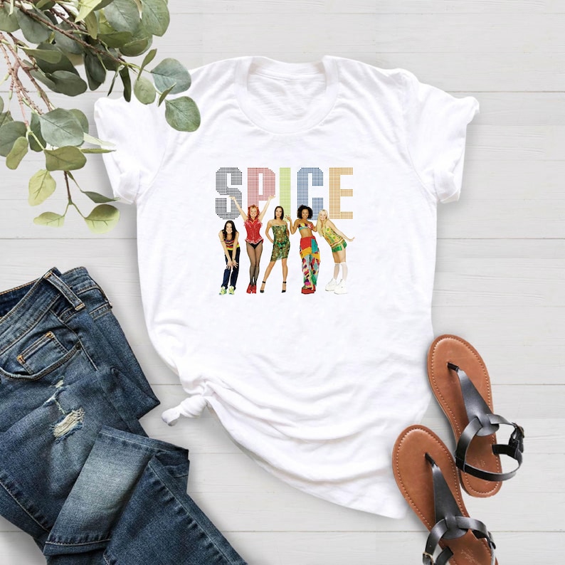 K&ouml;nnte beinhalten: Wei&szlig;es T-Shirt mit einem farbenfrohen Grafikdesign der Spice Girls und dem Wort "SPICE" dar&uuml;ber. Das Design zeigt die f&uuml;nf Mitglieder in verschiedenen Outfits. Das Shirt wird mit blauen Jeans und braunen Sandalen pr&auml;sentiert.