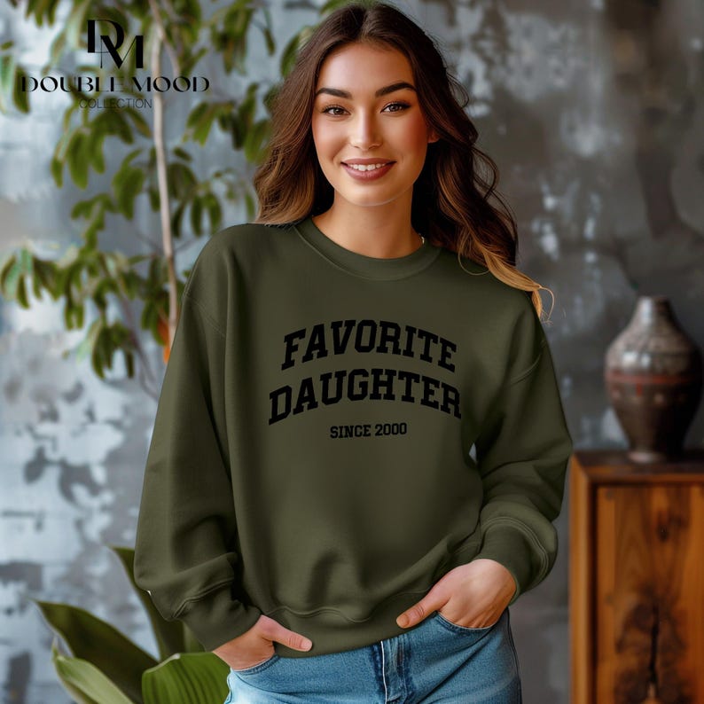 May include: An olive green sweatshirt with the text "FAVORITE DAUGHTER SINCE 2000" in black block letters. The sweatshirt is a crew neck style and is being worn by a person.