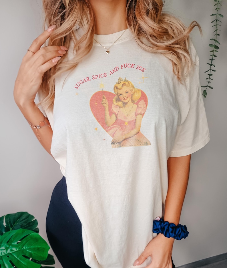 May include: Cream-colored t-shirt featuring a vintage-style illustration of a woman in a pink dress and crown inside a heart. The text "SUGAR, SPICE AND FUCK ICE" is arched above the heart. The shirt has short sleeves and a relaxed fit.