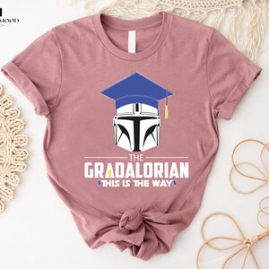 以下が含まれることがあります： ピンクのTシャツに、白いマンダロリアンヘルメットの上に青い卒業帽が乗っています。Tシャツには「The Gradalorian This is the Way」という文字がプリントされています。
