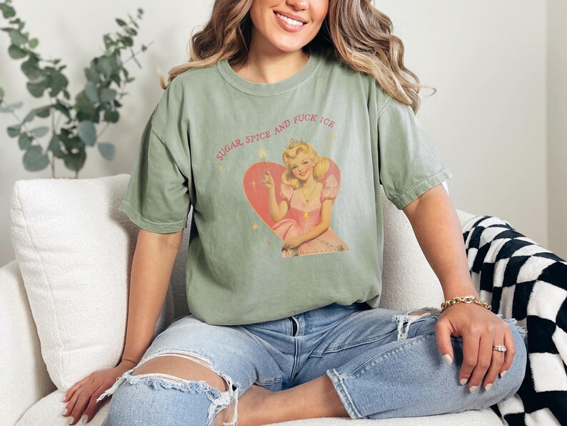 May include: Sage green t-shirt featuring a vintage-style illustration of a woman in a pink dress and crown inside a heart. The text "SUGAR SPICE AND FUCK ICE" is arched above the graphic. The shirt is made of a soft material.
