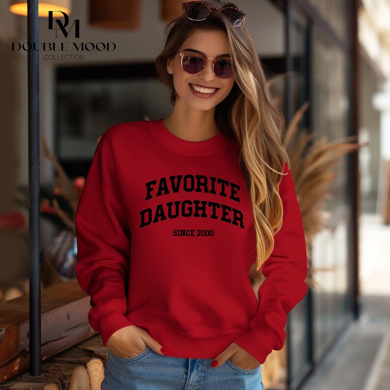 May include: A red sweatshirt with the text "FAVORITE DAUGHTER SINCE 2000" in black. The sweatshirt is a crew neck style and is made of a soft material. The Double Mood Collection logo is in the upper left corner.