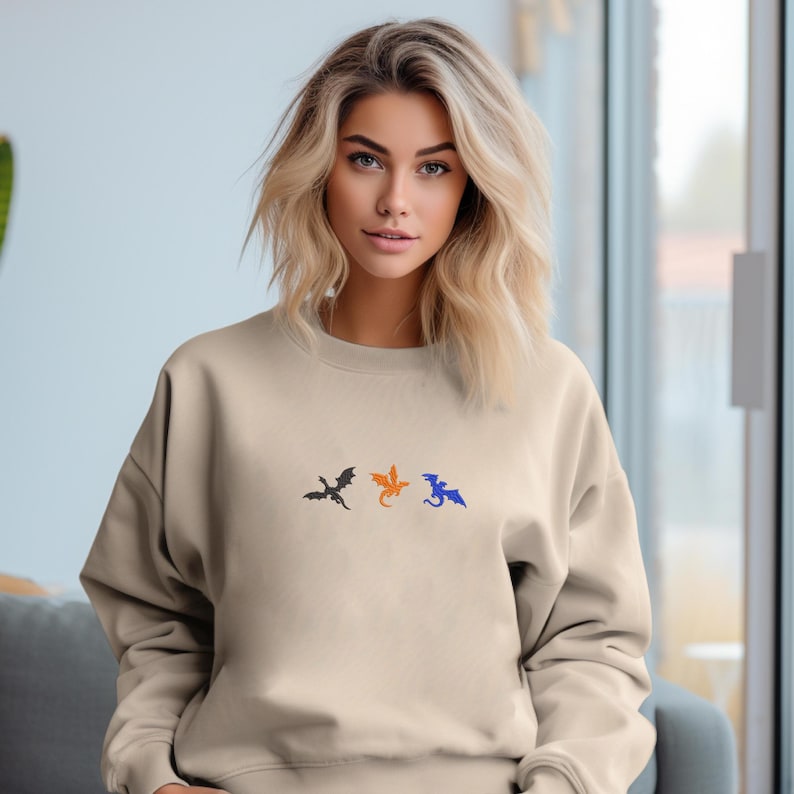 May include: Beige crewneck sweatshirt featuring three embroidered dragon silhouettes in black, orange, and blue. The dragons are arranged horizontally across the chest. The sweatshirt is a neutral color.
