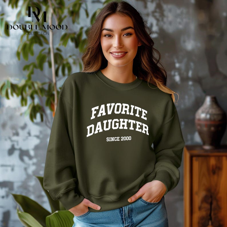 May include: An olive green sweatshirt with the text "FAVORITE DAUGHTER SINCE 2000" in white block letters. The sweatshirt is a crew neck style and is being worn by a person. The Double Mood Collection logo is in the upper left corner.