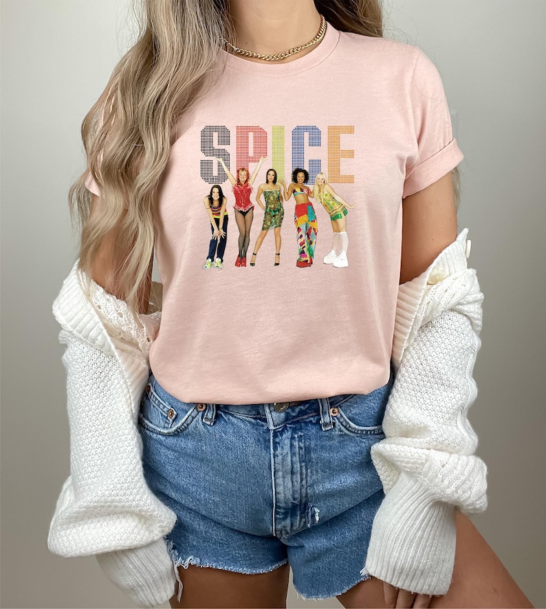 K&ouml;nnte beinhalten: Hellrosa T-Shirt mit dem Wort "SPICE" in einem bunten, pixeligen Schriftzug. Darunter befinden sich Illustrationen von f&uuml;nf Frauen in verschiedenen Outfits. Das T-Shirt hat kurze &Auml;rmel und einen Rundhalsausschnitt.