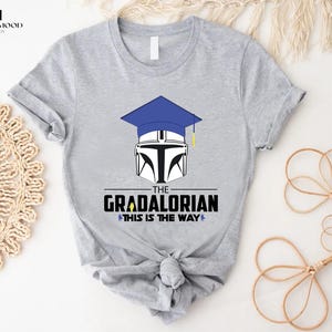 以下が含まれることがあります： グレーのTシャツに、青い卒業帽とマンダロリアンのヘルメットがプリントされています。Tシャツには「The Gradalorian This is the Way」という文字がプリントされています。