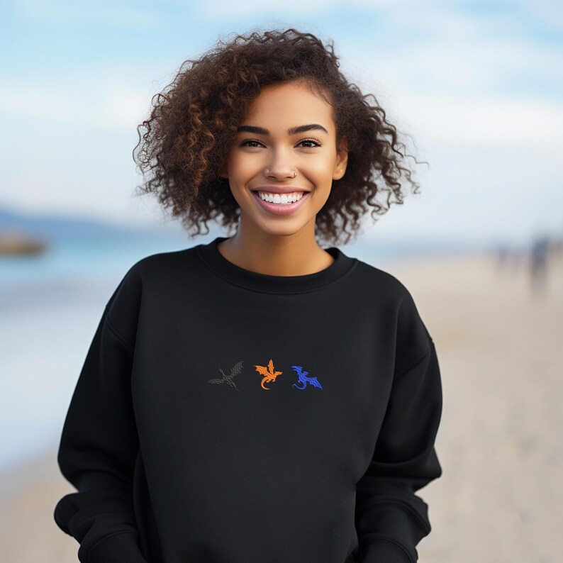 May include: Black sweatshirt featuring three embroidered dragons in gray, orange, and blue. The dragons are arranged horizontally across the chest. The sweatshirt is a solid black color.