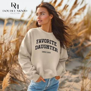 May include: A cream-colored sweatshirt with the text "FAVORITE DAUGHTER SINCE 2000" in black block letters. The sweatshirt is part of the Double Mood Collection. The model is wearing blue jeans and is standing outside.