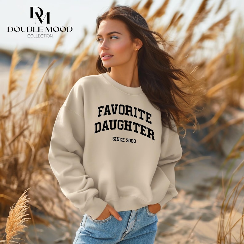 May include: A cream-colored sweatshirt with the text "FAVORITE DAUGHTER SINCE 2000" in black block letters. The sweatshirt is part of the Double Mood Collection. The model is wearing blue jeans and is standing outside.