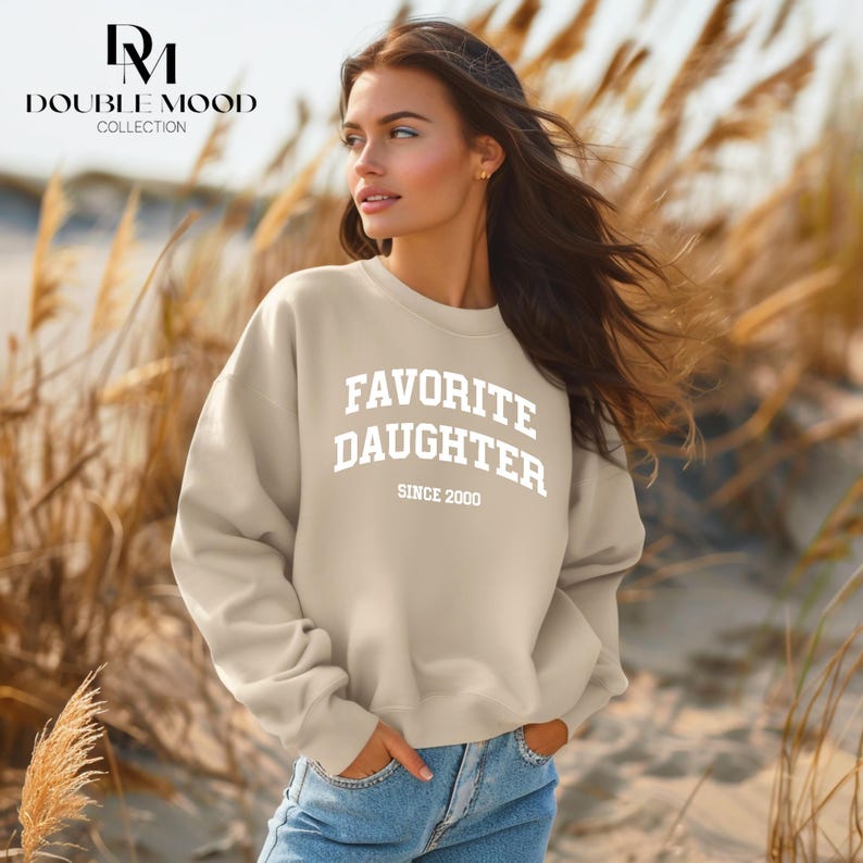 May include: A beige sweatshirt with the text "FAVORITE DAUGHTER SINCE 2000" in white. The sweatshirt is part of the Double Mood Collection. The model is wearing blue jeans and is standing in front of tall grass.