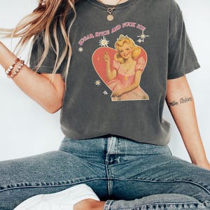 May include: Dark gray t-shirt with the text "SUGAR, SPICE AND FUCK ICE" above a vintage-style illustration of a woman in a pink dress and crown inside a heart. The shirt is paired with blue jeans and white sneakers.