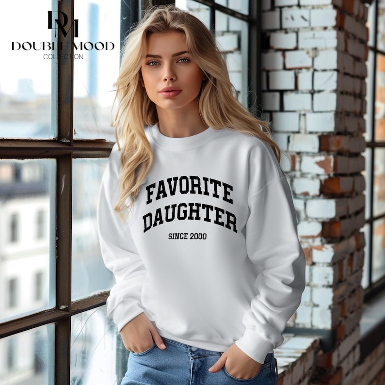 May include: White sweatshirt with the text "FAVORITE DAUGHTER SINCE 2000" in black block letters. The sweatshirt is long-sleeved and has a crew neck. The model is wearing blue jeans.