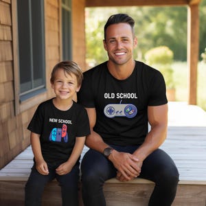 Op de afbeelding: Twee zwarte t-shirts met gaming-ontwerpen. E&eacute;n shirt heeft de tekst "OLD SCHOOL" met een controller-afbeelding, de andere "NEW SCHOOL" met een andere controller-afbeelding. Een man en een kind dragen de shirts.