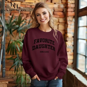 May include: Burgundy sweatshirt with the text "FAVORITE DAUGHTER SINCE 2000" in black. The sweatshirt is a crew neck style. The Double Mood Collection logo is in the upper left corner.