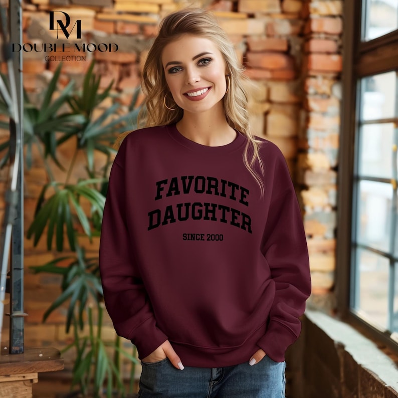 May include: Burgundy sweatshirt with the text "FAVORITE DAUGHTER SINCE 2000" in black. The sweatshirt is a crew neck style. The Double Mood Collection logo is in the upper left corner.