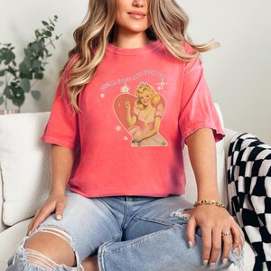 May include: Coral-colored t-shirt featuring a vintage-style graphic of a woman in a pink dress within a heart, with text above. The shirt is paired with blue jeans. The overall aesthetic is casual and retro.