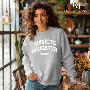 May include: A light gray sweatshirt with the text "FAVORITE DAUGHTER SINCE 2000" in white block letters. The sweatshirt is a crew neck style and is being worn by a person.