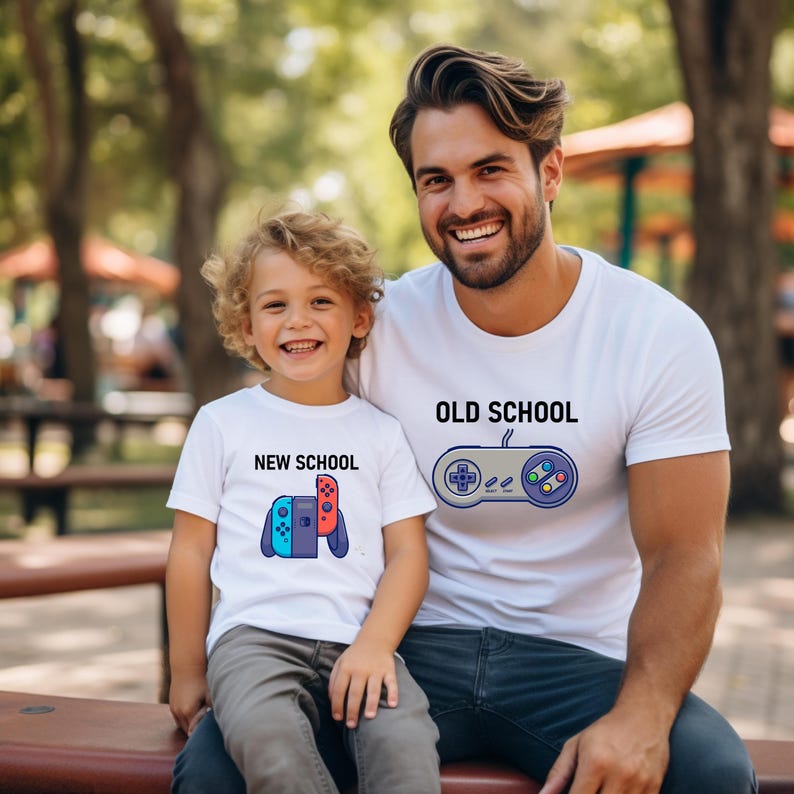 Op de afbeelding: Twee witte T-shirts met bijpassende ontwerpen. E&eacute;n shirt zegt "OLD SCHOOL" met een controller-afbeelding. De andere zegt "NEW SCHOOL" met een gamecontroller-afbeelding. De shirts worden gedragen door een man en een kind.
