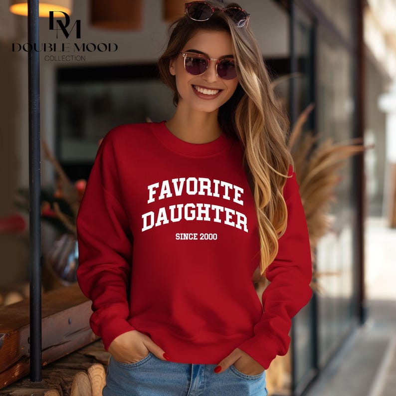 May include: A red sweatshirt with the text "FAVORITE DAUGHTER SINCE 2000" in white block letters. The sweatshirt is a crew neck style and is being worn.