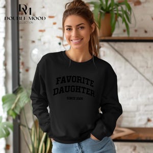 May include: Black sweatshirt with the text "FAVORITE DAUGHTER SINCE 2000" in a bold, block font. The sweatshirt is long-sleeved and has a crew neck. The Double Mood Collection logo is in the upper left corner.