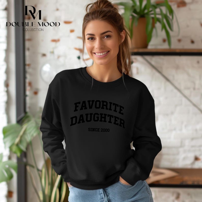 May include: Black sweatshirt with the text "FAVORITE DAUGHTER SINCE 2000" in a bold, block font. The sweatshirt is long-sleeved and has a crew neck. The Double Mood Collection logo is in the upper left corner.